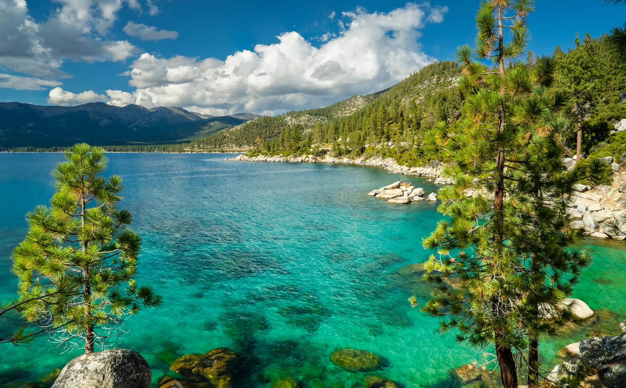 Clear turquoise water, rocky shores, pine trees, and mountains at Lake Tahoe.